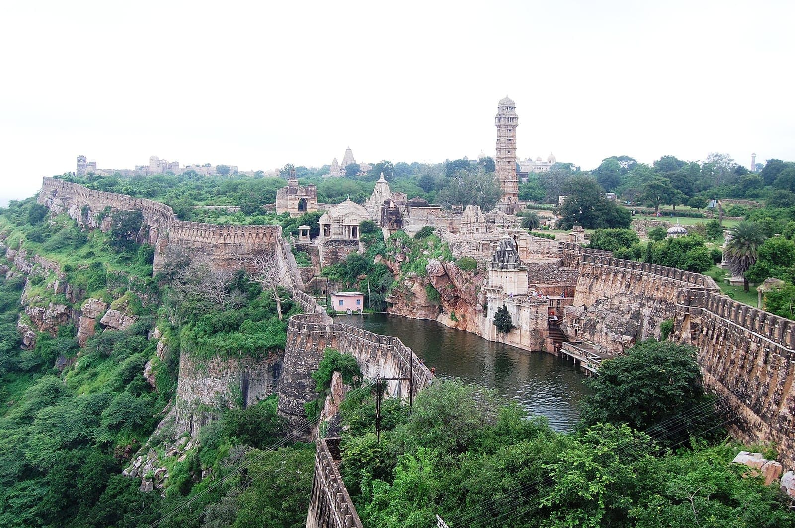 The walls of Chittorgarh Fort around a reservoir on a green hillside, with Vijay Stambha beyond