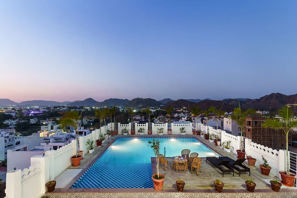 The rooftop pool glowing at dusk with the lights of Udaipur below the hills