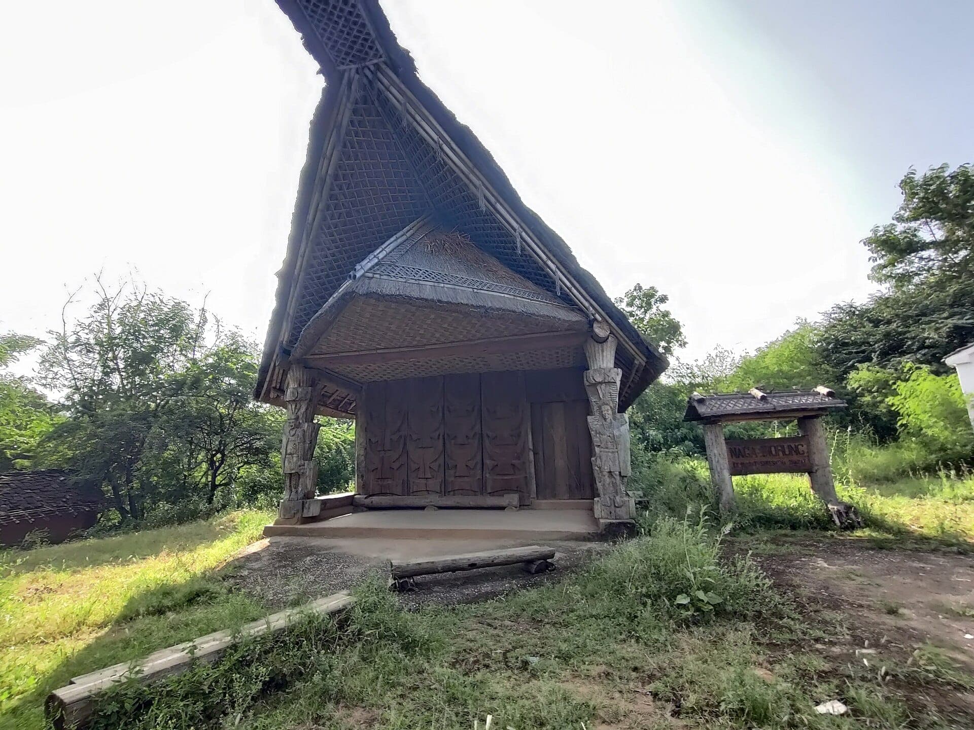 A carved wooden hut with a steep lattice-patterned roof among the trees at Shilpgram, Udaipur