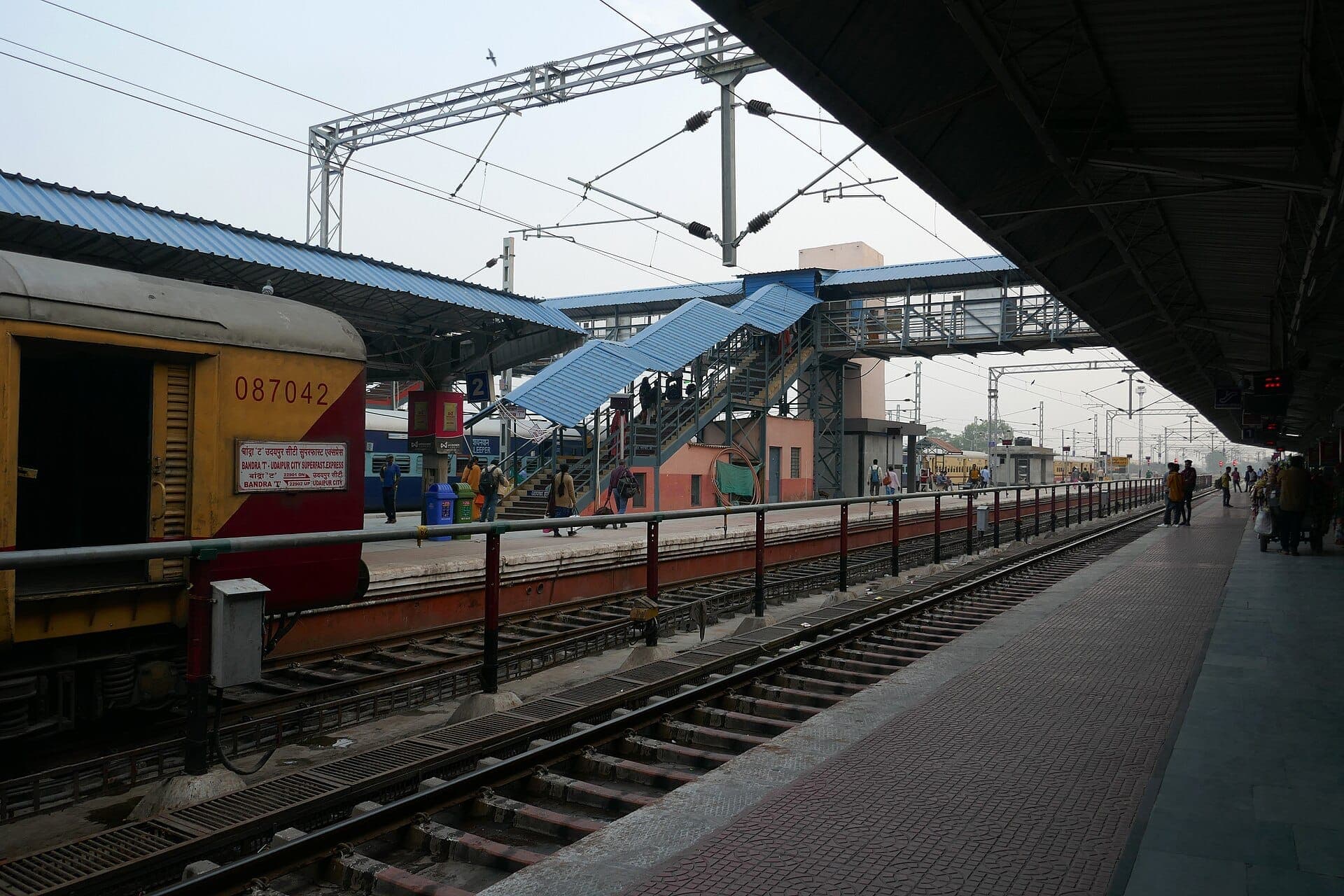 A train beside a platform at Udaipur City railway station, with the blue-roofed footbridge beyond