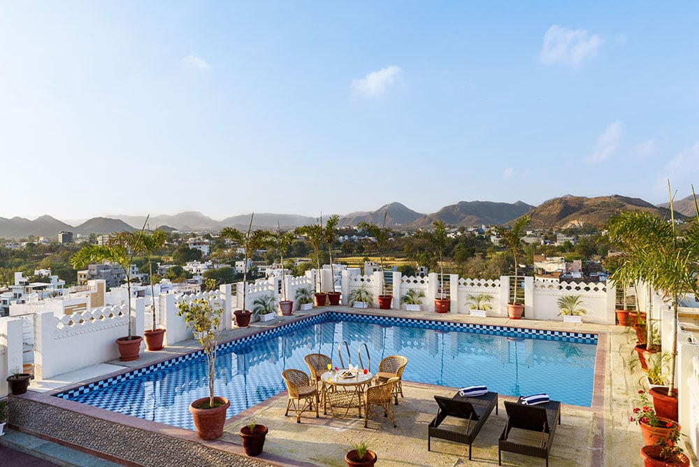Rooftop swimming pool with loungers, potted palms and the Aravalli hills in the distance