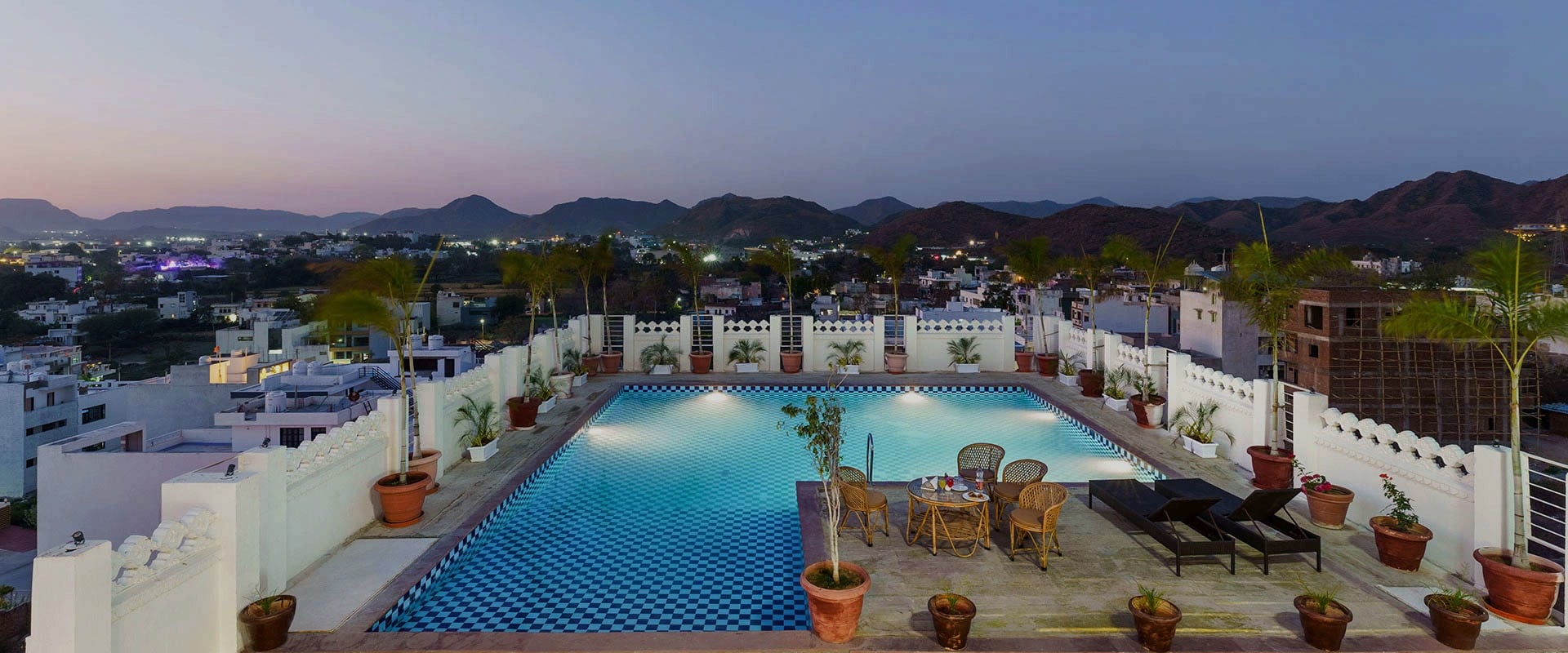 The rooftop swimming pool at dusk, lit from within, with the Aravalli hills beyond