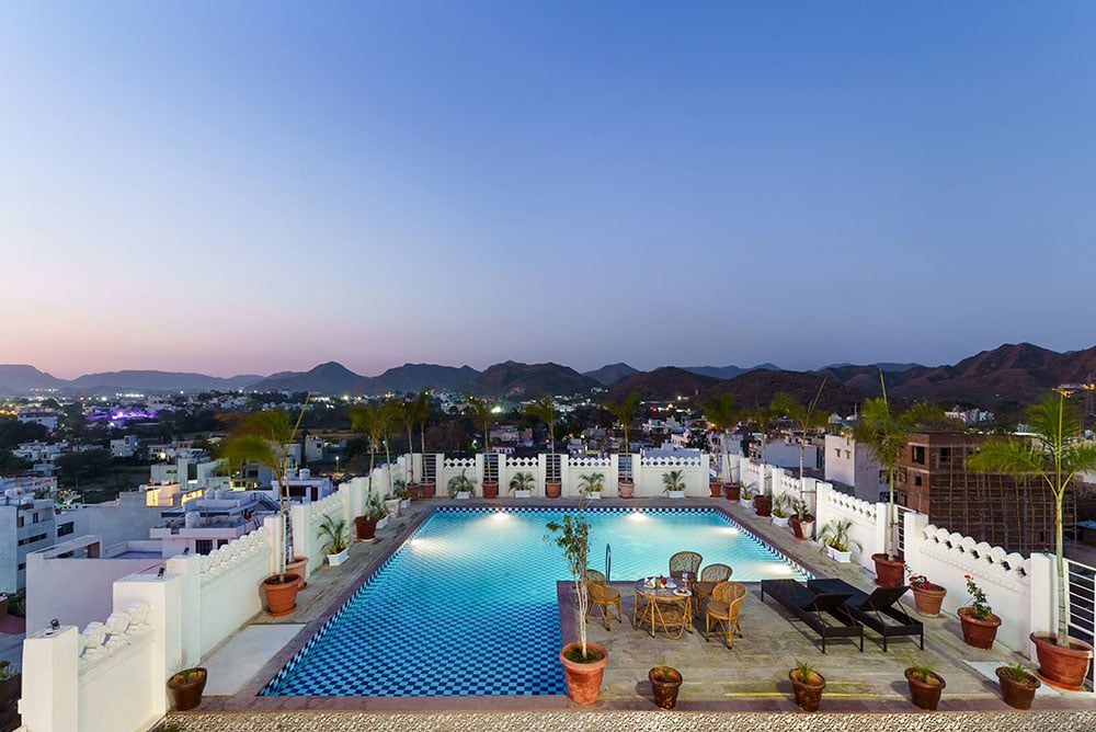 The rooftop pool glowing at dusk with the lights of Udaipur below the hills