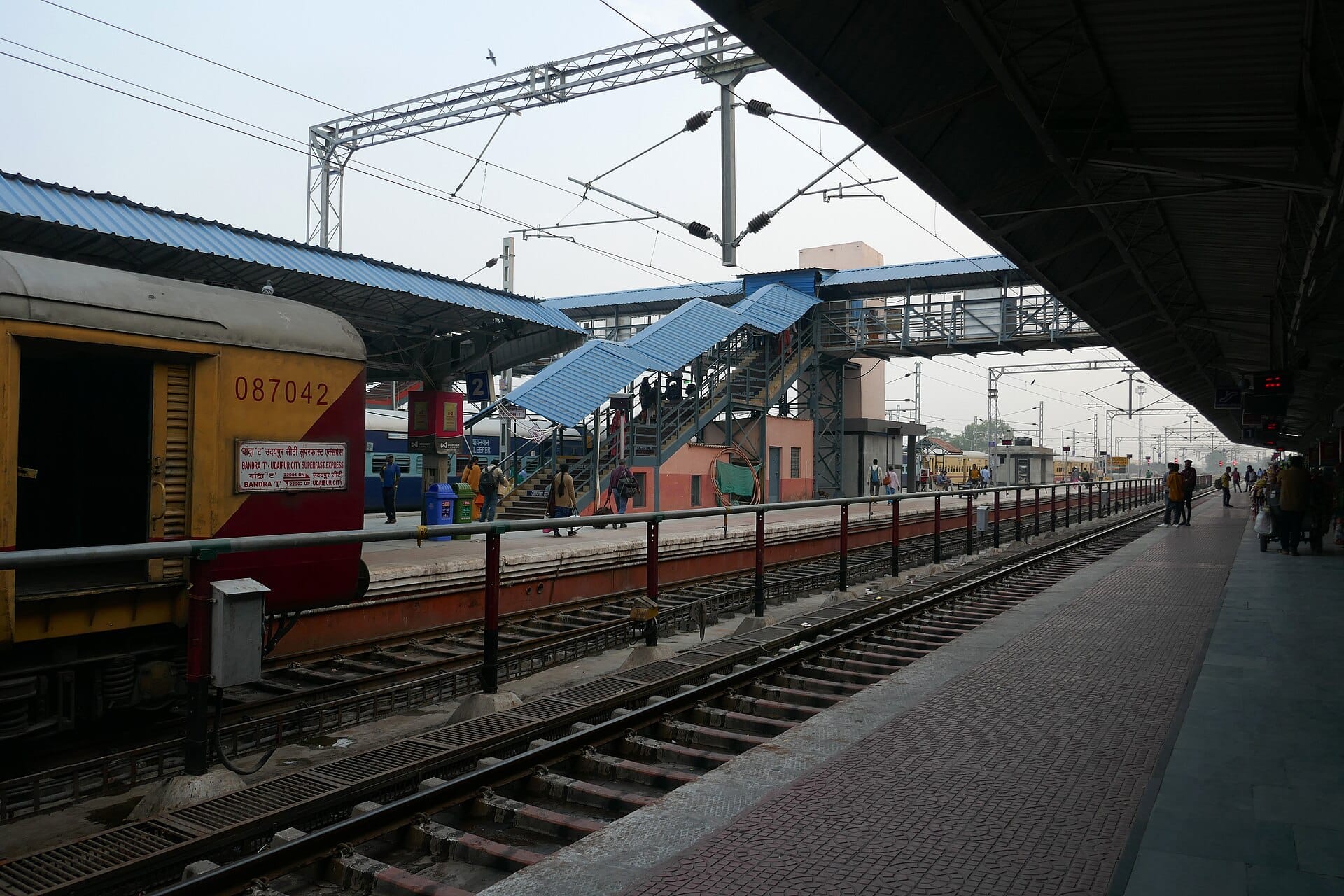 A train beside a platform at Udaipur City railway station, with the blue-roofed footbridge beyond